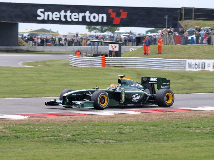 Pistonheads Snetterton - In the foreground, a green Formula 1 car is captured mid-turn on the racetrack, with its sleek design and vibrant color visible. The racetrack curves and we can see various spectators, some distant and others closer, watching the car from the stands and other designated areas. Behind the car, there are grandstands and areas that cater to the crowd to enjoy the race. In the background, there is a large advertisement with the words "Snippeton" visible, indicating the location or sponsor of the event. The grass surrounding the track is tidy and maintained.