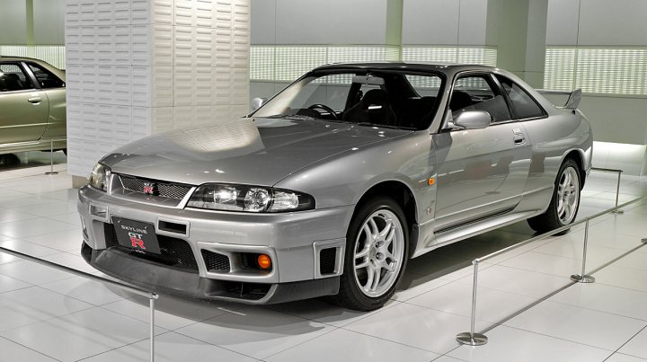 RE: Nissan Skyline R34 GT-R: Spotted - Page 6 - General Gassing - PistonHeads - The image displays a sleek silver sports car on display inside a car showroom. The car has a glossy finish and features deep lines and contours indicative of a luxury sports vehicle. It's an electric sports car, as revealed by its modern design and distinctive grille with an emblem, suggesting it might be a high-end model. The vehicle is parked behind a silver metal rod barrier, indicating that it is under showroom lighting, which highlights its reflective surface. In the background, a showroom interior can be partially seen, characterized by polished flooring and neutral color tones that complement the car's metallic hue.