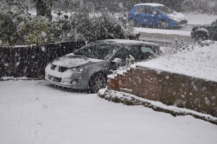 Wirral Lives Pistonheads - The image presents a wintry scene with a car parked next to a snow-covered street. The car, which appears to be silver, is covered in a thin layer of snow, suggesting that it has been there for some time. Another car, visible in the background, is driving on the road, partially covered in snow. Surrounding the parked car, there are trees dusted with the same snow, indicating a blanket of snow covering the entire area. The driveway of a nearby house is also visible, further emphasizing the wintry setting. The image captures the solitude of the parked car in the midst of a flurry.