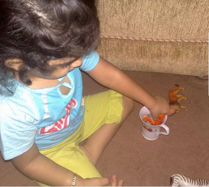 In the image, a young girl is caught in the middle of a playful moment. She is seated on the floor, her attire consisting of a blue t-shirt and yellow shorts. In her right hand, she holds a small toy rocket. Simultaneously, her left hand is about to crush an orange toy car. The background of the image is a wooden floor, providing a casual and comfortable setting for her imaginative playtime.