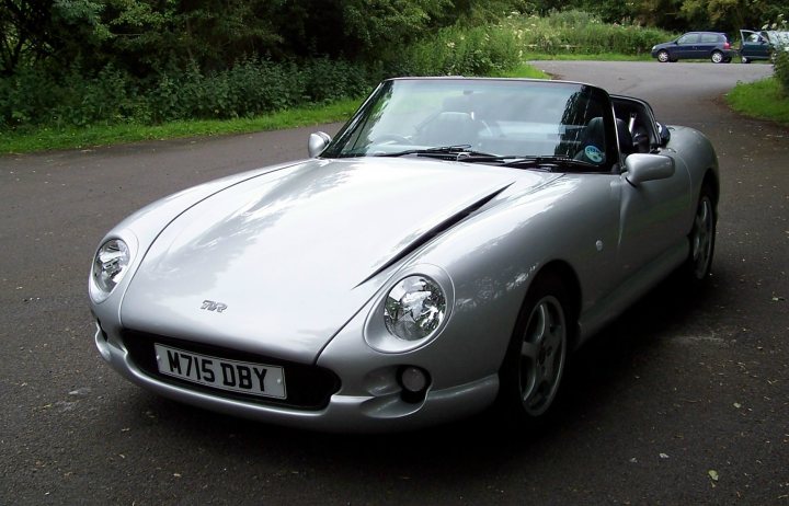 Change Big Bulbs Wheelarch Pistonheads Hole - The image shows a silver sports car parked on an asphalt surface. The vehicle is a convertible with its top down, revealing two seats in the interior. In the background, there are natural surroundings with green foliage. Further back, there's a glimpse of another car and a road curving to the right. The overall atmosphere of the image is serene and sunny, suggesting a pleasant driving day.