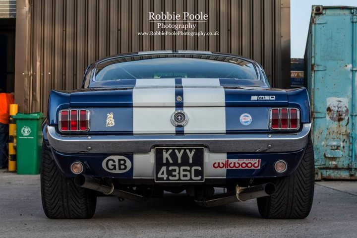 The GT Cafe - the ace cafe of the north - Aug meet - Page 1 - North West - PistonHeads - The image showcases a classic Mustang car, painted in striking stripes of blue and white. The vehicle is parked in a location that appears industrial, with brown metallic structures and a bright blue dumpster in the background. The Mustang has a custom license plate that reads "KY436C" and several badges and a cylinder emblem on the trunk, suggesting it is a modified and possibly a performance model. The photo is credited to Robbie Poole, of Robbie Poole Photography, indicating he may be the owner of the car or simply the photographer.