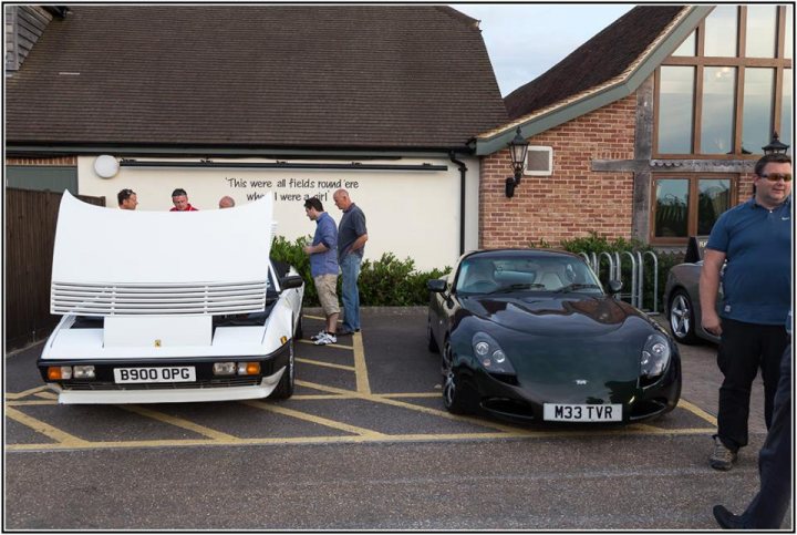 July TVR + Friends meet - Page 1 - Thames Valley & Surrey - PistonHeads - The image portrays a man with blue hair standing near two cars, a white classic and a sleek black sports car, parked adjacent to each other in front of a building. The building has a well-maintained front garden and a sign that says, "THE WATKIN YARD, THE WATKIN ROUND AND THE WATKIN FIELD WERE ALL PARK ROUND." There is a sense of nostalgia as well as a welcoming environment. The overall scene suggests a peaceful day at a historical or preserved area.