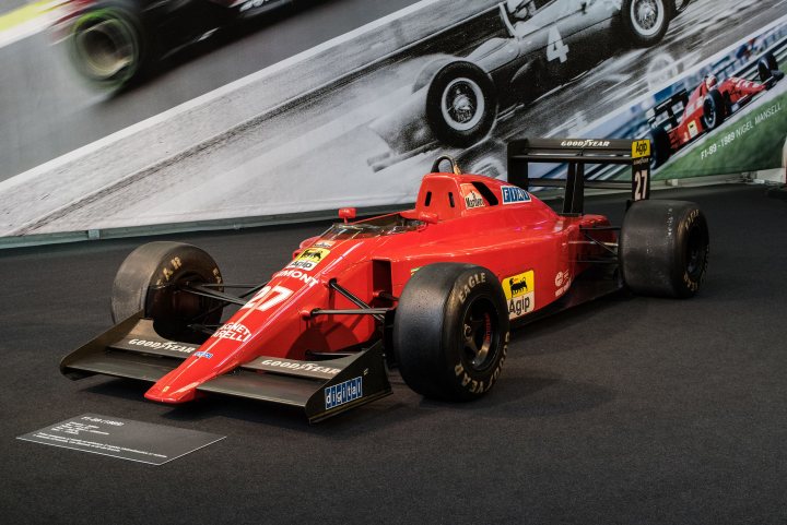 Favourite F1 cars 1980 onwards  - Page 12 - Formula 1 - PistonHeads UK - The image showcases a vibrant red racing car exhibited on a black surface. The car is adorned with various sponsor logos and has a prominent front wing. In the background, there's a sign displaying images of racing cars, suggesting that this scene could be part of an exhibition or event related to motorsport. A blurred image of a person is visible at the top right corner, possibly indicating someone observing the car. The overall setting seems to be indoors, and the lighting creates a dramatic effect on the car's shiny surface.