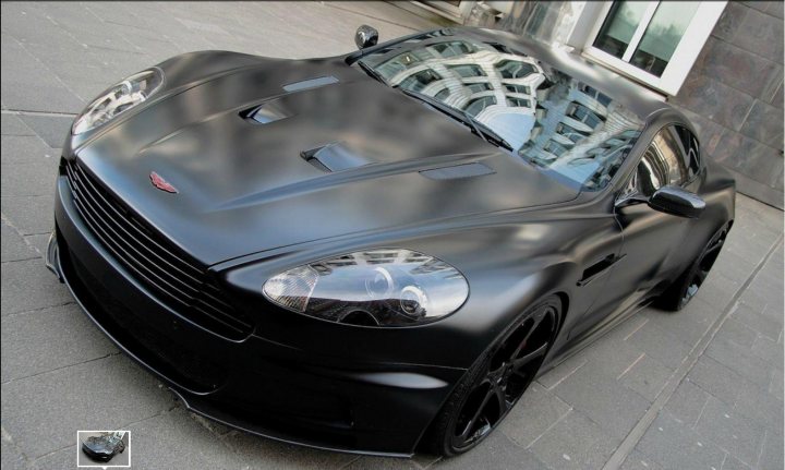 Which colour vehicle wrap? - Page 1 - Aston Martin - PistonHeads - The image showcases a sleek black sports car, parked in a serene setting. The contrast between the car's glossy black exterior and the bricks of the building next to it is striking. The vehicle, characterized by its two doors and modern design, sits on black rims, complementing its overall dark hue. The calm, spacious street where the car is parked adds a sense of tranquility to the scene.