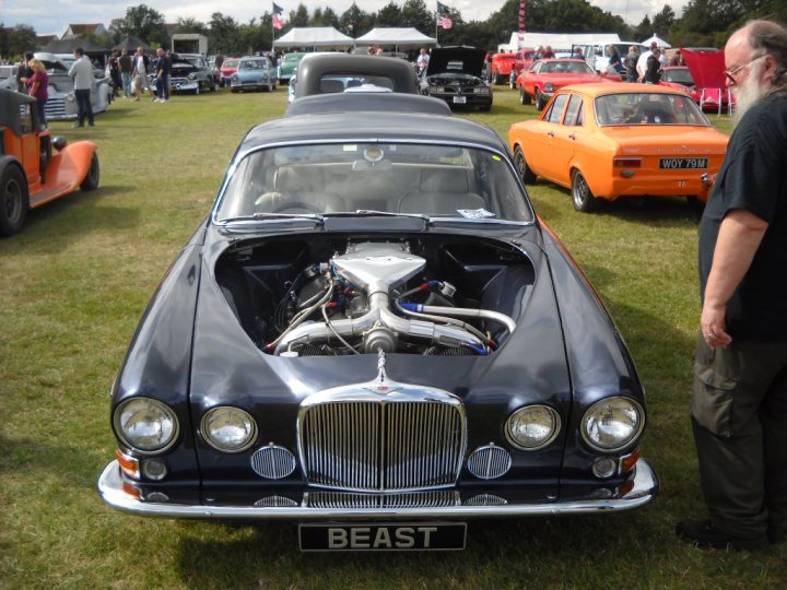 Anyone going to the NASC 40th Nats at Trinity Park? - Page 1 - East Anglia - PistonHeads - The image depicts an outdoor car show on a grassy field. The central focus is on a classic blue car with a visible silver engine. There are several other vehicles parked around the field, including older cars and trucks. In the foreground, a man in sunglasses and a black shirt is standing beside the classic blue car, possibly admiring or discussing it with others. The sky is overcast, and there are flags and tents set up in the background, suggesting a larger event or gathering.