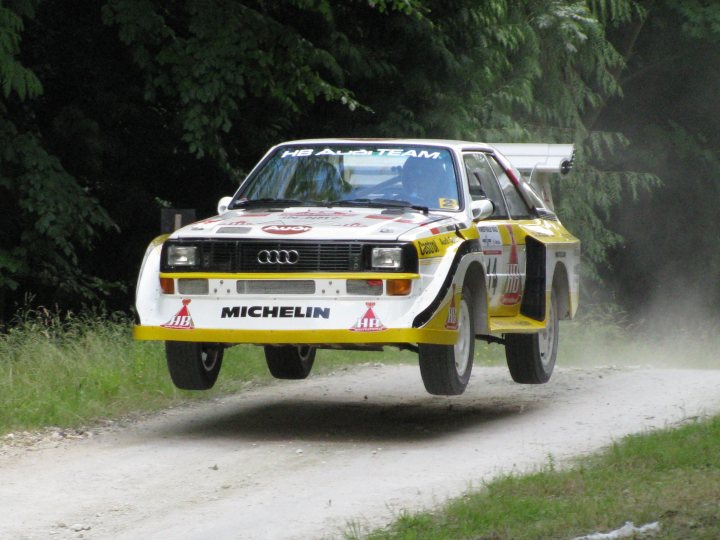 Pistonheads - The image captures a dramatic scene on a gravel road where a white rally car, bearing the number 18 and the name "Heuer Alicat," is performing stunts. The car is in the midst of a jump, raising high into the air with its black and yellow tires against the backdrop of trees. The back of the car prominently displays the logo "MICHELIN," indicating the type of tires used in the event. The suspension of the car is stretched out as it's caught in mid-air, showcasing the power and finesse involved in this daring sport.