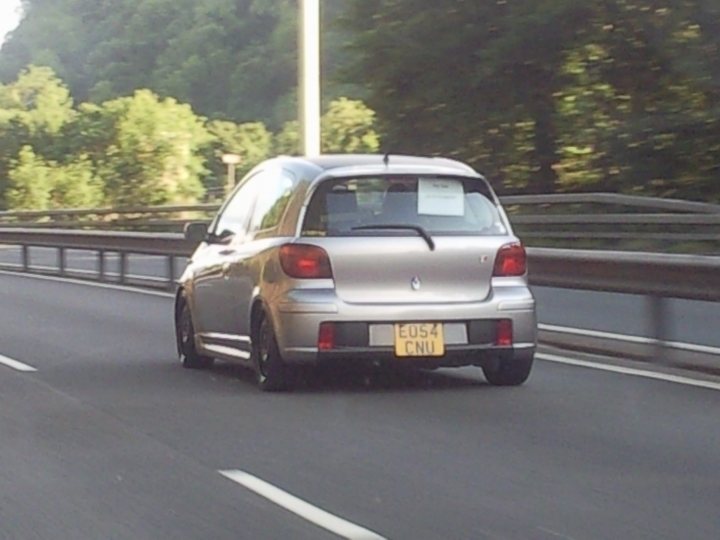 hi guys im new here...... - Page 1 - South Wales - PistonHeads - The image depicts a silver car in motion on a highway. The car is a compact hatchback, a common type of vehicle used for urban commuting. The license plate is visible, showing the registration number EUS4 CNI. The car is driving on the right side of the road, adhering to the standard road rules of the country. The surrounding area appears to be a wooded hillside with green trees, suggesting an area of natural beauty or a highway cutting through a forested region.