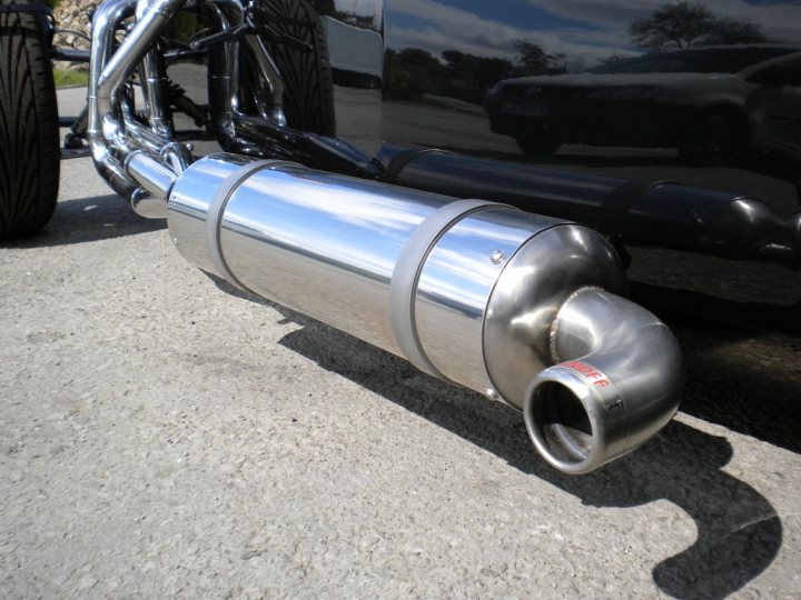 Anyone have experience with WunOff exhausts - Page 1 - Caterham - PistonHeads - The image shows a close-up of the exhaust pipe of a vehicle. The pipe appears to be made of a shiny, metallic material, likely chrome or stainless steel. Its design indicates that it's for a high-performance vehicle, possibly one associated with racing or luxury. The chrome finish reflects the surrounding light, which suggests it might have been taken during the day. The pipe is positioned on the ground, under the vehicle it is attached to. There is also a reflection of a tree on the surface it rests on, which provides contrast and further emphasizes the shiny and reflective nature of the pipe.