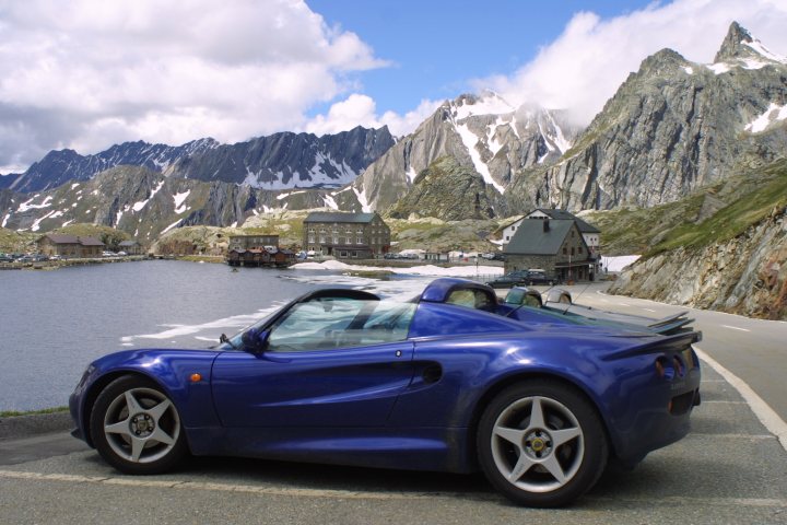 The big Elise/Exige picture thread - Page 1 - Elise/Exige/Europa/340R - PistonHeads - The image captures a serene outdoor setting, dominated by a blue sports car parked on the side of a road. The car is sleek and shiny, suggesting it's in good condition. The road on which the car is parked seems almost abandoned, with the steep mountains on one side and a tranquil lake on the other, populated only by a few boats bobbing gently. The sky above is partly cloudy, adding to the dramatic beauty of the scene. The color palette of the image is dominated by cool tones, from the blue of the car and lake to the gray of the mountains and the muted colors of the sky, contributing to a calm and tranquil vibe.