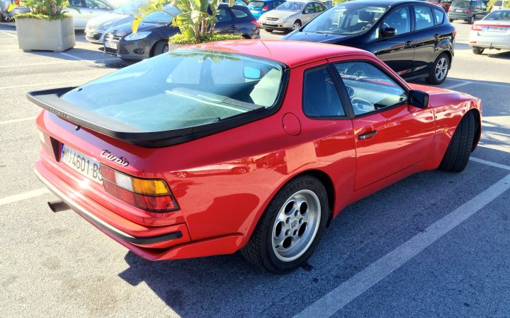 My first venture into Porsche land... - Page 1 - Front Engined Porsches - PistonHeads - The image features a vibrant red classic sports car parked on an asphalt surface within a parking lot. The car is facing left, and its windows are down. The vehicle's rear window raises above the roofline, a common characteristic of BMW sports cars. In the background, there are a few other vehicles and clusters of trees, indicating the photo might be taken in early spring or late summer due to the full bloom of trees.