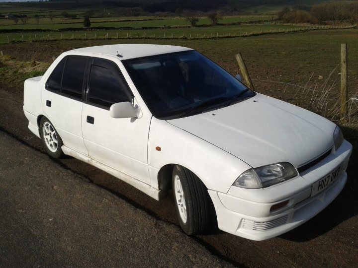 A small white car parked in a parking lot - Pistonheads - The image features an older model white car parked on a paved road. It's positioned at an angle to the camera, revealing its passenger side and front part of the vehicle. The car appears to have some bird droppings on its side, indicating it might have been parked for a while. The setting is in what seems to be a rural area with grassy fields and a wire fence just behind the car. There are no people or other vehicles in sight, suggesting it might be a quiet or sparsely populated location.