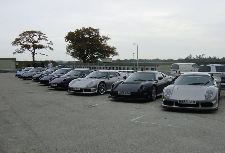 A group of cars parked on the side of the road - Pistonheads - The image showcases a parking lot teeming with life, occupied with a variety of cars and trucks, their colors ranging from the pristine white to the sleek black, and the rugged gray. Some are parked in neat rows, others are scattered, creating a dynamic and bustling scene. Amidst this mechanical mingling, a solitary tree stands tall, providing a touch of nature amidst the concrete. The sky overhead is a canvas of overcast clouds, casting a soft, diffused light over the scene. Despite the gloomy weather, there's a sense of movement and activity in this image, as if capturing a moment in the daily hustle and bustle of life.