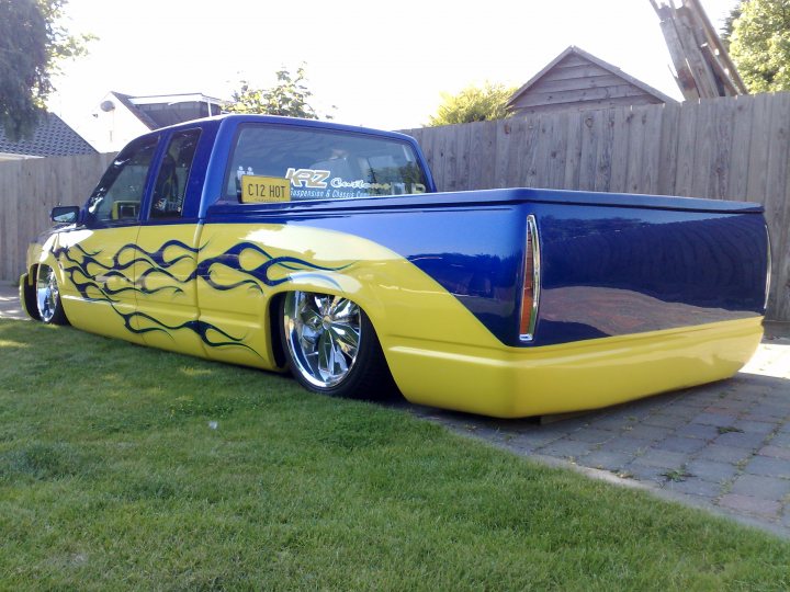 Chevy Pistonheads Truck - The image showcases a striking yellow pickup truck in a residential area. The vehicle bears a vibrant blue flame design along its side, adding an element of custom style to its appearance. Parked alongside a wooden fence and lush grass, the truck is the main focus of the image, giving a sense of a well-kept home environment. The truck's size is accentuated by a particularly large spare tire mounted in the bed, adding to its utilitarian appeal.