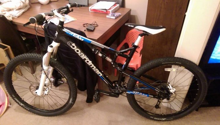 The "Show off your bike" thread! - Page 379 - Pedal Powered - PistonHeads - In the image, a high-angle shot of a blue and black mountain bike is captured, parked indoors on a carpeted floor. The bike's position in the room suggests it's ready to be ridden. In the background, various items of disarray are scattered around, including a book and a remote control, indicating a lived-in space. To the right, there's a glimpse of a large TV screen mounted on the wall. The overall scene conveys a casual, home-like setting with the bike as the central focus.