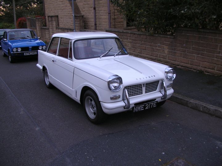 Herald Nmee White Pistonheads Lincs Area - The image features a vintage white car parked on the side of a street. The car has a distinctive design, reminiscent of the 1960s, with round headlights and black detailing. It's a two-door vintage car with the license plate securely on the front. In the background, there's another car farther down the road, indicating that this photo was taken in an urban setting. The focus of the image is the vintage car, capturing its details and presence on the road.