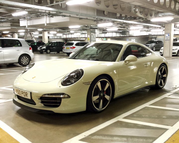 Supercars spotted, some rarities (vol 6) - Page 292 - General Gassing - PistonHeads - This image shows a bright white Porsche sports car parked in a multi-story parking garage. The car is sleek with a design that suggest speed and performance. Various other cars can be seen in the background, parked in different orientations, which indicates a public parking area. The garage itself has a high ceiling with visible ductwork, and there are cars parked in both the upper and lower sections, highlighting the multi-story nature of the space. The lighting is artificial, with round, bright lights that provide ample illumination to the area.