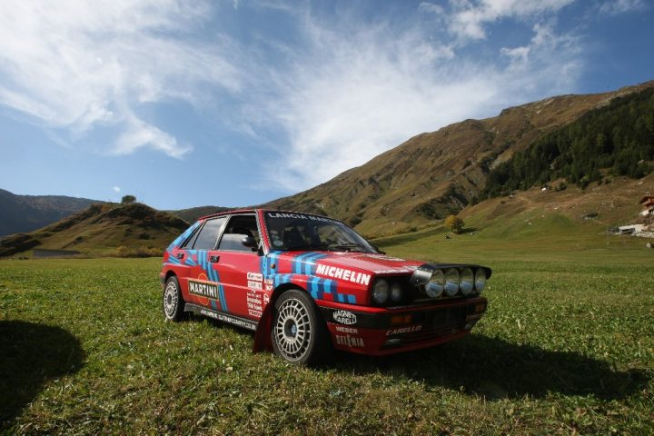 Some half decent pics - Page 1 - North West - PistonHeads - The image presents a vibrant scene in a grassy field dotted with mountains in the background. A striking red car, adorned with a striking blue and black pattern, adds a pop of color to the tranquil landscape. The car's detailing suggests it could be a vintage or rally car, enhancing the adventurous spirit of the setting. The vehicle is angled towards the viewer, giving a sense of depth and inviting them into the scene to join in the journey.