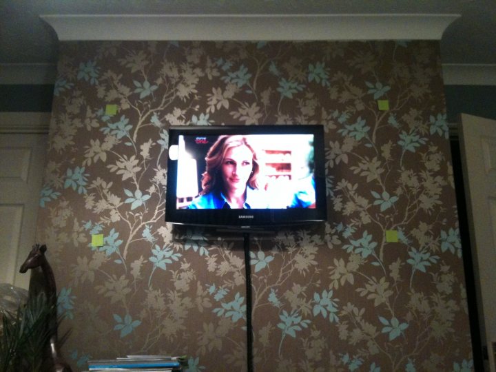 Pistonheads Size Recommendation - The image displays a television set mounted on a wall with wallpaper. The television screen is displaying what appears to be a woman talking, possibly on a news program based on the visible graphic and text. To the left of the television, air fresheners are placed, and a partially visible plant is barely visible. The overall setting suggests a living room or a similar room designed for television viewing.