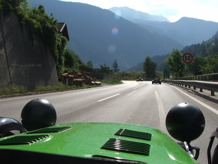 Not enough pictures on this forum - Page 8 - Caterham - PistonHeads - This image captures a moment on a winding mountain road. At the forefront, a green vehicle with a distinctive black front and bright headlights is driving away from the viewer. A red and white speed limit sign stands on the right side of the road, indicating a limit of 60. The road is surrounded by lush greenery and a gray stone wall is visible on the left side, possibly indicating a boundary or safety measure. The background is dominated by the majestic presence of mountains, suggesting that the driver is in a mountainous region.