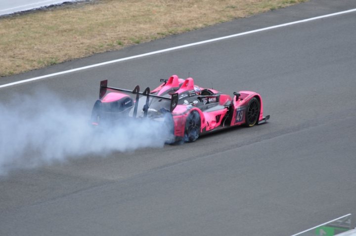 Le Mans 2011 - Page 1 - Vipers - PistonHeads - The image captures a thrilling scene on a race track. A car, with a distinctive pink and black body, is the main focus. It's in motion, with its rear smoke stack creating a dramatic outburst of smoke against the open sky. The car is leaning to the right, indicating high speed or aggressive maneuvering, which is typical in a racing context. The background reveals a well-maintained track with clear markings, suggesting a professional racing environment. The colors, dynamic action, and the adrenaline-inducing atmosphere all contribute to the sense of excitement and speed.