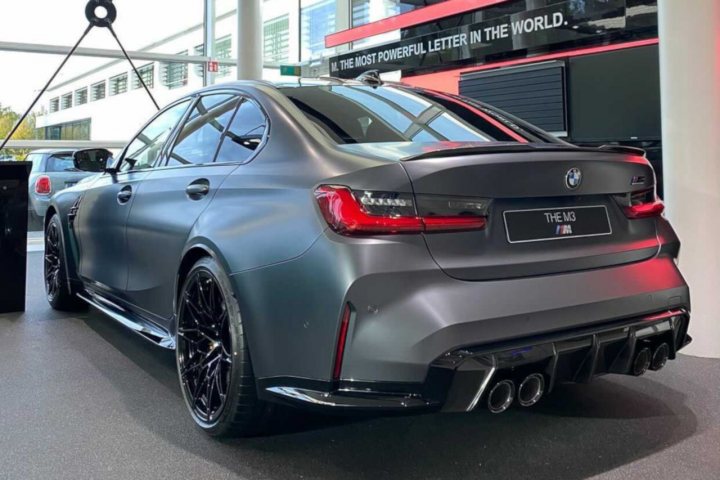 The image shows a customized BMW car on display, likely at a car show or similar event. The vehicle has a striking matte grey finish that contrasts with the black wheels. Its bodywork features custom add-ons, including what appears to be an aftermarket kit that includes side skirts and additional vents. The interior of the car is not visible in this image. In the background, there are other cars, suggesting this might be a large auto show or exhibition. There are also people present, but their faces are not clearly distinguishable due to the angle of the photo. The setting appears to be an indoor venue with artificial lighting, and the overall ambiance is one of sophistication and automotive culture.