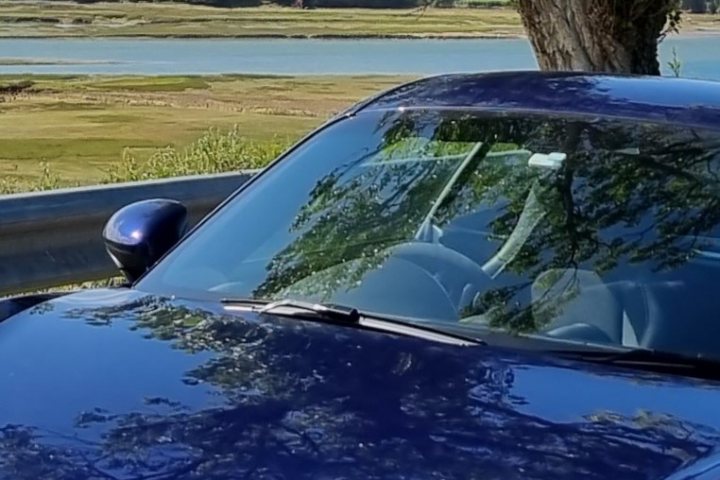 Pistonheads - This image depicts a car parked by the side of a road, which appears to be near a body of water. The car is blue and has a sunroof open. In the background, there's a grassy area with trees, suggesting a rural or suburban setting. Above the car, the sky is visible with clouds and what seems like a mountain in the distance. There are no people or other vehicles visible in this image. The style of the photo suggests it was taken during daylight under natural lighting conditions.