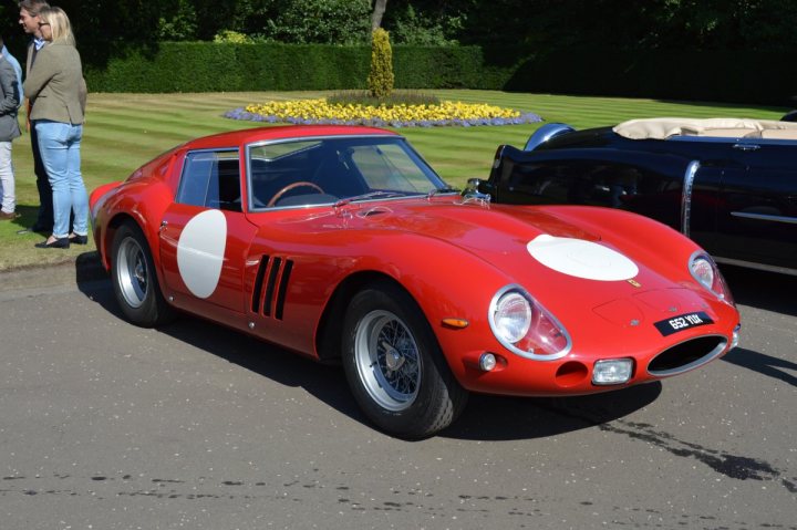 A red car with a surfboard on top of it - Pistonheads - This image features a classic red sports car parked on a paved surface. The car is adorned with two white racer-style stripes on its side, adding to its vintage appearance. The license plate is clearly visible and proudly states "5529". The setting appears to be a garden or a park, with neatly trimmed hedges and trees in the background, indicating an outdoor event or gathering.