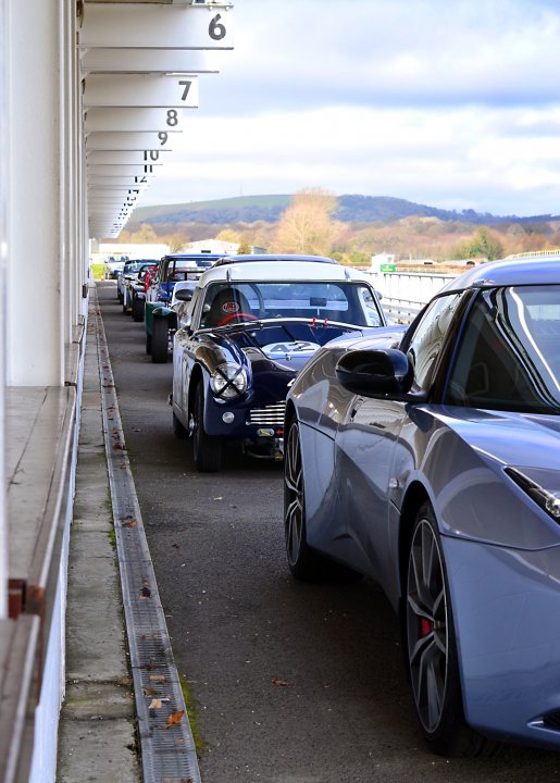 Robert Barrie Track Day 24th Feb Goodwood - photos - Page 1 - South Coast - PistonHeads - The image presents a row of luxury cars neatly parked under a covered structure. The cars vary in color, with hues ranging from sleek dark tones to vibrant metallic shades, reflecting the soft light. The structure appears to be part of an indoor parking facility, as suggested by the grid-like markers lining the viewing area. The environment outside the structure is blurred but reveals a lush, green landscape under a bright, sunny sky.