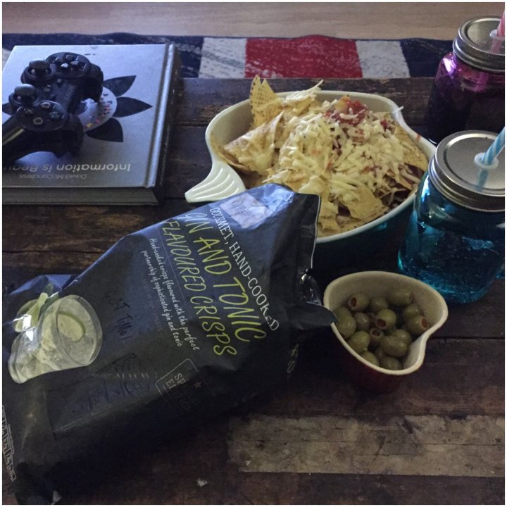 ALDI Food Worth Trying?  - Page 74 - Food, Drink & Restaurants - PistonHeads - The image showcases a casual, cozy scene on a wooden table. There is a large bag of Doritos with the snack "Flaming Hot Mattatos" prominently displayed on it, a plastic container filled with tortilla chips, a small bowl filled with green olives, and a jar filled with tonic-flavored garnishes. Next to these items are a blue straw and a drink, suggesting someone was enjoying a beverage. The table also holds a hardcover book and a pair of sunglasses, adding a touch of familiarity to the setting. Nearby on the floor, there's a book and a remote control, hinting at a relaxed and leisurely atmosphere.