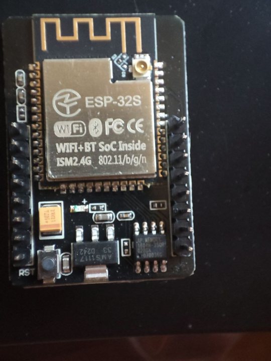 This is a photograph of an electronic device, specifically a small white circuit board with various components and labels. The circuit board features an EEPROM chip at its center with the text "ESP32" on it, indicating it's based on the ESP32 microcontroller by Espressif Systems. Around the EEPROM chip are other chips and connections, suggesting it is part of a larger electronic system. The circuit board is attached to a surface, likely a table or desk, which is partially visible in the image. The background behind the circuit board appears blurry, focusing attention on the device itself.