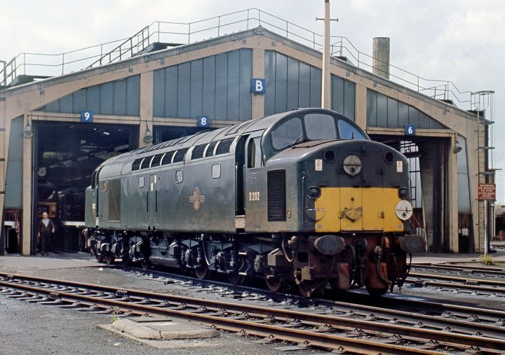 Loco sheds and other railway buildings.... - Page 16 - Boats, Planes & Trains - PistonHeads UK - This is a color photograph of an old-style diesel locomotive parked outside of what appears to be a train station or maintenance facility. The train has a prominent black and yellow livery, which suggests it may be a preserved or historical model rather than in active service. There are no visible people in the immediate vicinity of the train, but there is a person partially visible on the right side of the image. In the background, you can see a building with what looks like a garage door and various other vehicles, including another locomotive or possibly a diesel engine, to the left. The setting suggests an industrial environment, and the lighting indicates it might be daytime.