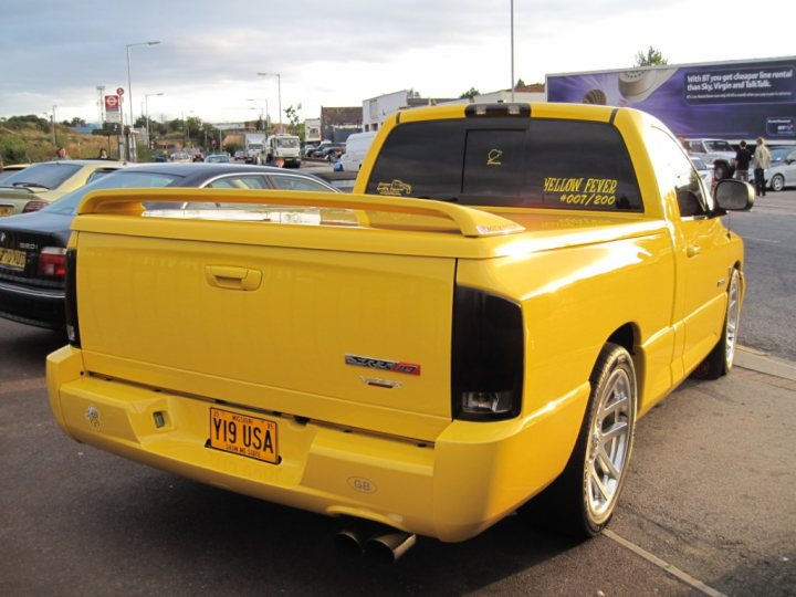 Yellow Pistonheads Fever - The image showcases a vibrant yellow Dodge Ram truck with tinted windows, parked in a lot. The back of the truck features a tow hitch, equipped to handle various tasks. The truck is parked next to another black truck and a silver car, indicating a shared parking space. In the background, the lot extends to a striking purple billboard, adding a pop of color to the scene.