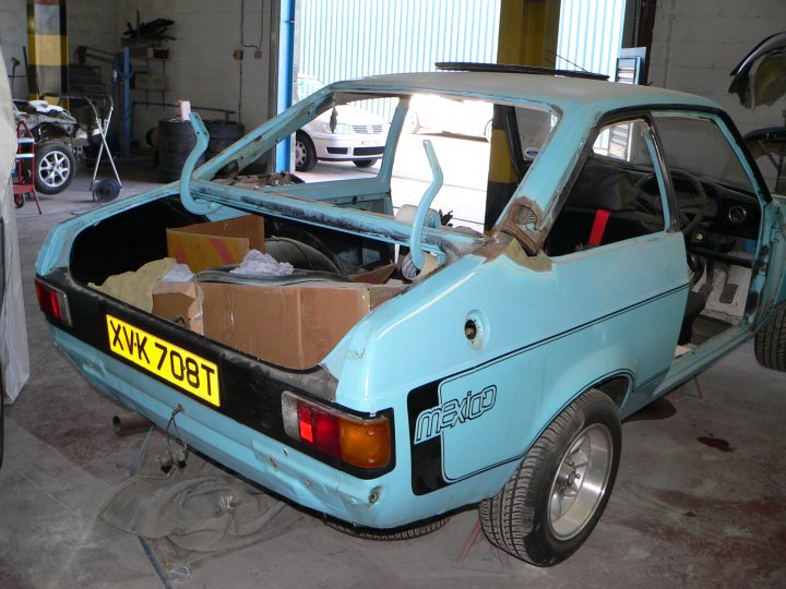 Escort Pistonheads Popular - This image features a vintage blue car parked inside a garage building. The car's hatch is open, revealing items stored within, such as tools, clothes, and household goods. There are two spare tires and a luggage rack attached to the car. The vehicle's roof appears to be removed, suggesting that the interior is being cleaned or packed. The garage environment is somewhat disorganized, with a variety of objects and vehicles scattered around the car.