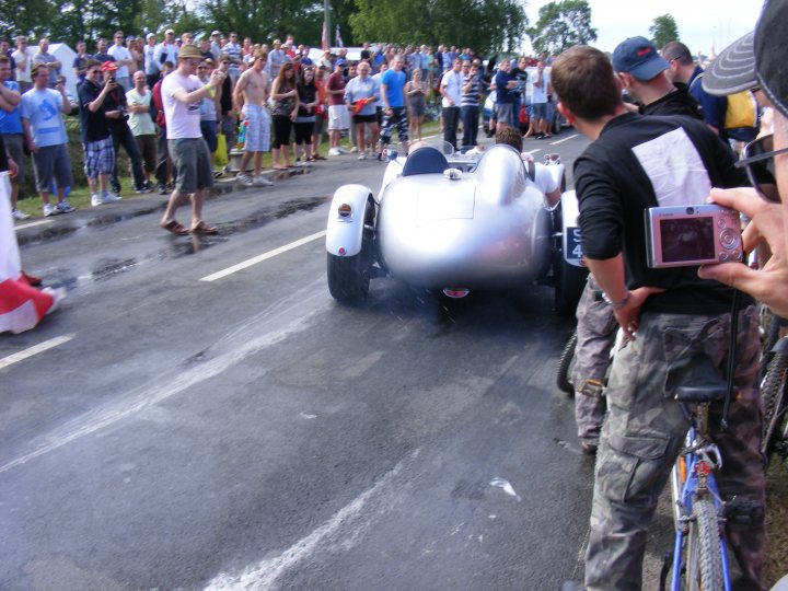 Pistonheads Classics - The image captures an exciting moment at an event where a silverberg with an aggressive profile is being ridden by a man. The car is dense, suggesting speed or perhaps afterburners, and appears to be performing or maneuvering on a road. In the background, a large crowd is visible, their attention riveted on the action in front of them. Two individuals stand out from the crowd, one is holding a camera, possibly filming the event, and the other is holding a cell phone, likely to take photos or record the spectacle. The atmosphere seems to be one of anticipation and thrill, with the crowd eagerly watching the car's movements.