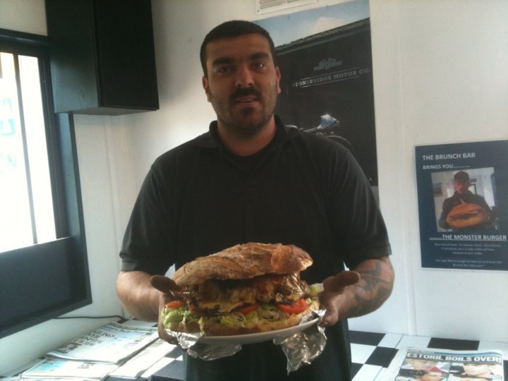 Jaw Ache Fancy Pistonheads - In the image, a man is standing in a room that appears to be set up for breakfast. He is wearing a polo shirt and is holding a large sandwich which is significantly larger than a standard burger. The man's expression is one of surprise or shock. Behind him, there's a large picture hanging on the wall, possibly related to the breakfast menu or theme. The floor beneath him has a black and white checkered pattern and there are what look like magazines or newspapers lying around, perhaps suggesting a leisurely morning breakfast.