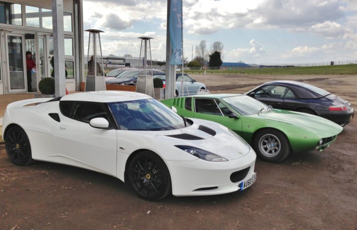 Some nice snaps - Page 1 - Evora - PistonHeads - The image features a white sports car parked prominently on the right, showcasing its sleek design and black rims. Adjacent to the white sports car is a vibrant green sports car, adding a pop of color to the scene. Further back, a black car is visible. The cars are parked in a lot that seems to be a car dealership, indicated by the presence of the dealership entrance and other cars displayed for sale. The setting suggests a day out, possibly a shopping or car exploration trip.