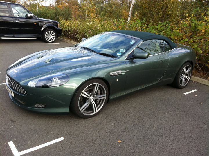 The non-black-non-grey-non-silver thread - Page 2 - Aston Martin - PistonHeads - The image captures a moment in a parking lot where a green Aston Martin reaching deep into its convertible design makes a striking contrast with a neatly parked black SUV. The Aston Martin, sitting on the driver's side of a parking space, features the iconic "Aston Martin" branding on the hood and side, along with some stickers used to fix a front license plate, which is missing. The moment is set against the backdrop of a calm day, with trees twinkling subtly in the distance.