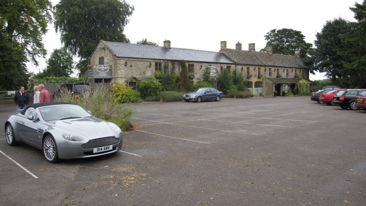 Pistonheads - The image shows a parking lot with several parked cars in front of a large, old building with a pointed roof. The building appears well-maintained, suggesting it might be hosting an event or is a commercial or public establishment. A sports car with the license plate "LE 48 AVY" is prominently parked near the center of the image, in the foreground. There are no people visible in the image. The sky is overcast, indicating it might be a cool, cloudy day. The lot is paved and devoid of any noticeable activity.