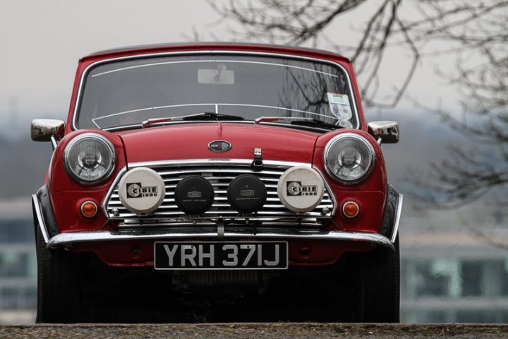 Brooklands Mini Day - Page 1 - Classic Minis - PistonHeads - The image shows a vintage red car with a distinctive silver grill featuring the insignia of a luxury brand. The car is parked on a road, with an overcast sky visible in the background. The vehicle's design, which includes a sizable chrome grill, is indicative of older models, suggesting a classic or retro aesthetic. The car has white round bar-like designs around the ends of the bumper, and a dark plate attached near the middle of the car. The lighting in the image appears to be natural, with diffused lighting indicating an overcast day.