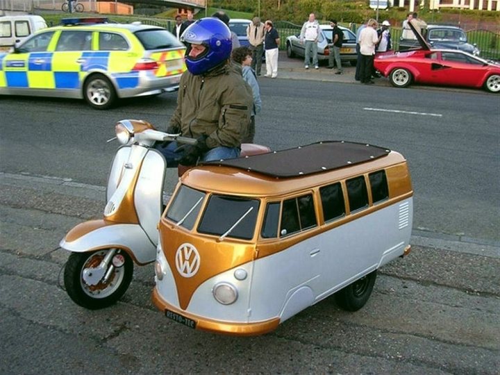 The scooter caravan - Page 1 - Tents, Caravans & Motorhomes - PistonHeads - The image captures a whimsical scene on a road where a man is driving a small, orange Volkswagen bus. The VW bus is noticeably small but is painted in a vibrant shade of orange, which sets it apart on the road. The man, wearing a helmet, is seated in the bus, seemingly in motion, avoiding obstacles like parked cars on the street. The street seems to be lined with various vehicles, and a few people can be spotted in the background.