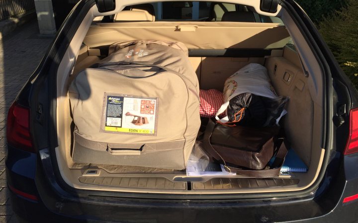 Quick pitching tent required - Page 1 - Tents, Caravans & Motorhomes - PistonHeads - The image shows the back of a dark-colored car filled with various items. At the top, there's a tan-colored duffel bag, which seems rich in texture. Behind the duffel bag, there's a white plastic bag partially contained within the trunk. Beside the duffel bag, a brown wooden handbag is placed, lying flat. The handbag appears to be made of leather and features a handle at the top and a shoulder strap. The car is parked in an area that is partially visible, with some greenery and a concrete surface in the background. The presence of these personal effects suggests that someone is either in the process of moving or preparing for a trip.