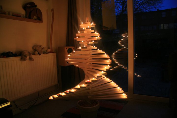 Thought I would make my Christmas Tree this year. (Pics). - Page 1 - Homes, Gardens and DIY - PistonHeads - The image depicts a cozy living room scene at night. The centerpiece is a large, uniquely designed Christmas tree made from white wooden planks. The tree is adorned with rows of lit candles that cast a warm glow on the surroundings. Flanking the tree on the left is a shelf with several stuffed animals, possibly teddy bears, arranged neatly and illuminated by soft, indirect light. To the right, there's an open window, silhouetting a house outside. The ceiling is painted in a dark color, contrasting with the curtains that are drawn. The overall atmosphere suggests a snug and inviting space, perfect for the festive season.