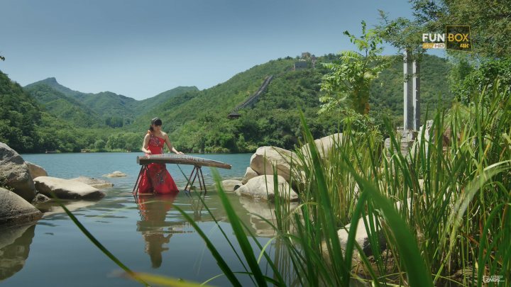 Two people sitting on a bench in the middle of a lake - The image depicts a woman in a stunning natural setting. She is seated on a large wooden board resting on the water, preparing to paddle, which appears to be a stand-up paddleboard. The waterway is surrounded by lush greenery, suggesting a serene and untouched environment. In the distance, there's a sky line hinting at the presence of a city beyond the forest-like area. Across the water, there's a structure with stairs, possibly a part of a larger natural park or resort. This setting could be a part of an adventure resort or a similar location that promotes outdoor activities and tourism.