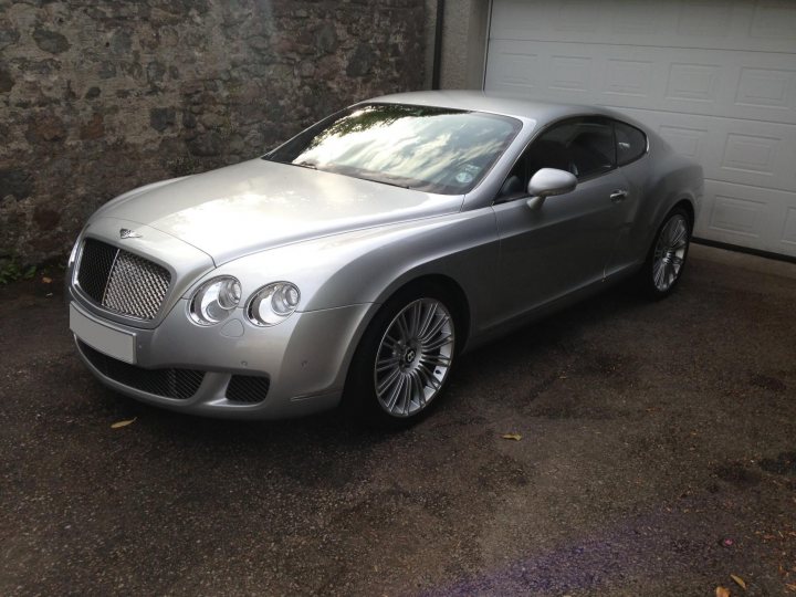 I’ve put my football boots on & bought a Bentley GT Speed - Page 1 - Rolls Royce & Bentley - PistonHeads - The image features a silver-colored luxury car, specifically a Bentley, parked in a driveway alongside a garage door. The car has a sleek and polished appearance, with several design elements typical of high-end models, including distinctive matrix grille, headlamps, and a prominent front fender with a rounded shape. The car is positioned at an angle that offers a clear view of the front and side profiles, and it's placed on what appears to be a black, asphalt-like driveway, which contrasts with the car's lighter color. The presence of the garage door indicates that this might be a residential setting.