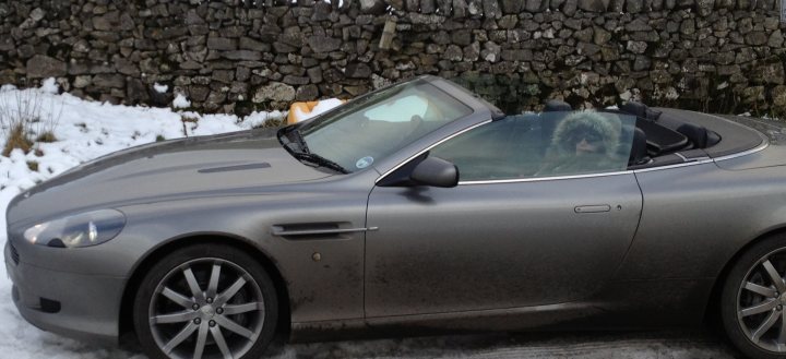 Using an Aston Martin in this weather? - Page 5 - Aston Martin - PistonHeads - The image captures a silver, two-door luxury car parked on a snowy road. The car's top is down, revealing a single person sitting inside. The person, donned in a full winter coat and hat, is looking out the back window. The backdrop of the image is a rocky wall, suggesting a rural location. The overall atmosphere is chilly and serene, typical of a winter's day.