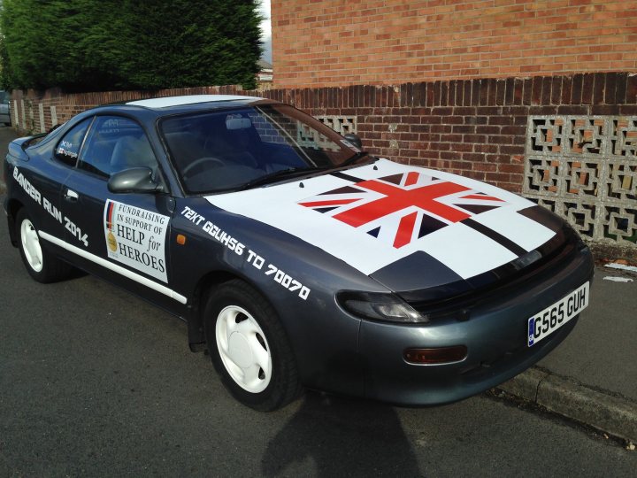 7 Countries 4 days & a banger - 'Help For Heroes' - Page 1 - South West - PistonHeads - The image features a black sports car parked on the side of a road. The car is adorned with a large white and red design, resembling the British flag, on its side. There are phone numbers and texts painted in white on the fender and the rear side of the car. The background of the image shows a dark red brick wall with uniform rectangular holes for what appears to be vine cuttings.