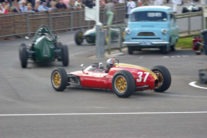 2012 Goodwood Revival Meeting Photo Gallery - Page 1 - Goodwood Events - PistonHeads - The image captures an exciting scene from a vintage race car event. In the foreground, a red race car with the number 37 and the driver's number 12 zooms through a curve toward the left of the frame. Two green race cars are also visible, competing for the lead. The scene is set in a parking lot, which serves as a racing track for these nostalgic machines, surrounded by spectators eagerly watching the action-packed race. The old car in the background adds to the vintage atmosphere of the event.