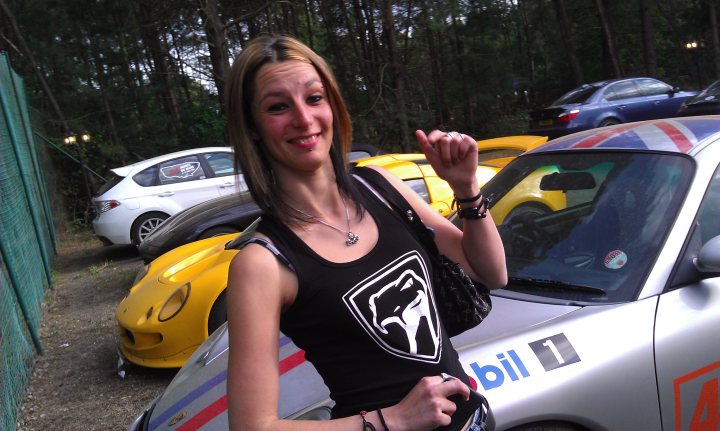 Le Mans 2011 - Page 1 - Vipers - PistonHeads - This image captures a cheerful scene in a parking lot. At the center of the image is a young woman, sporting a chic black top adorned with a white logo. She strikes a confident pose, with her hand on her hip and her other hand casually holding a delightful pink bag. Her radiant smile adds to the overall jovial mood of the image.

The parking lot is filled with an array of cars. A silver car, boasting a striking blue logo, and a yellow car emerge as the main attractions in this scene. They are parked in the backdrop, creating a beautiful contrast with the central figures.

Adding a touch of green to the scene are trees visible in the background, providing a natural backdrop to this vibrant tableau. The location appears to be during the day, under what seems to be a clear sky.