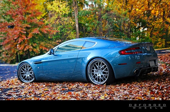 Car Porn - Page 1 - Aston Martin - PistonHeads - The image showcases a blue Aston Martin sports car parked on a leaf-strewn path. The car's sleek design is highlighted against the backdrop of a fall-colored forest, with the vehicle facing towards the right side of the frame. The color of the car contrasts beautifully with the rich hues of the autumn foliage. This evocative scene appears to be available for stock photo use, as indicated by the text "d2forgedwheels" present in the image.